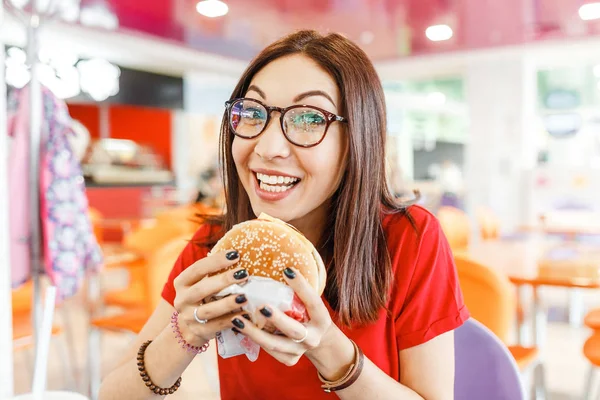 Szczęśliwy zdrowa kobieta siedzi pomieszczeniu gastronomicznej i jedzenie pyszne hamburger, koncepcja nowoczesnego posiłek — Zdjęcie stockowe