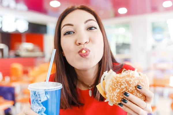 Щаслива молода жінка з гамбургером, содовою і картоплею фрі у ресторані швидкого харчування — стокове фото