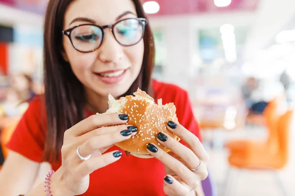 Счастливая здоровая женщина сидит в помещении фуд-корт и есть вкусный гамбургер, современная концепция еды — стоковое фото