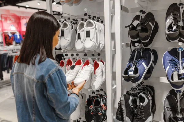 happy young woman customer choosing pair of sneakers shoes and buying it in clothes store, casual shopping concept