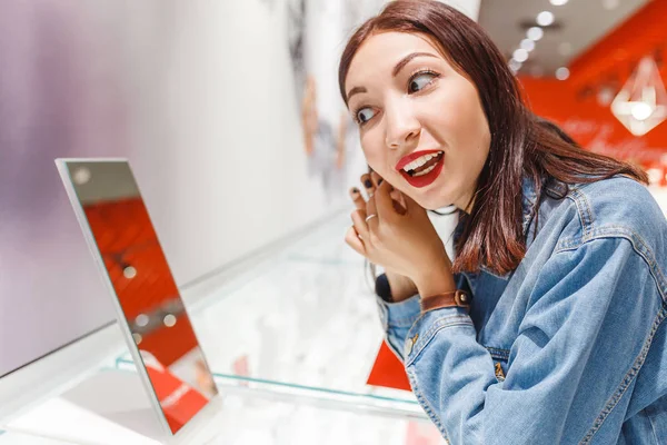 Hermosa mujer joven probándose pendientes y mirando al espejo en la joyería, concepto de compras — Foto de Stock