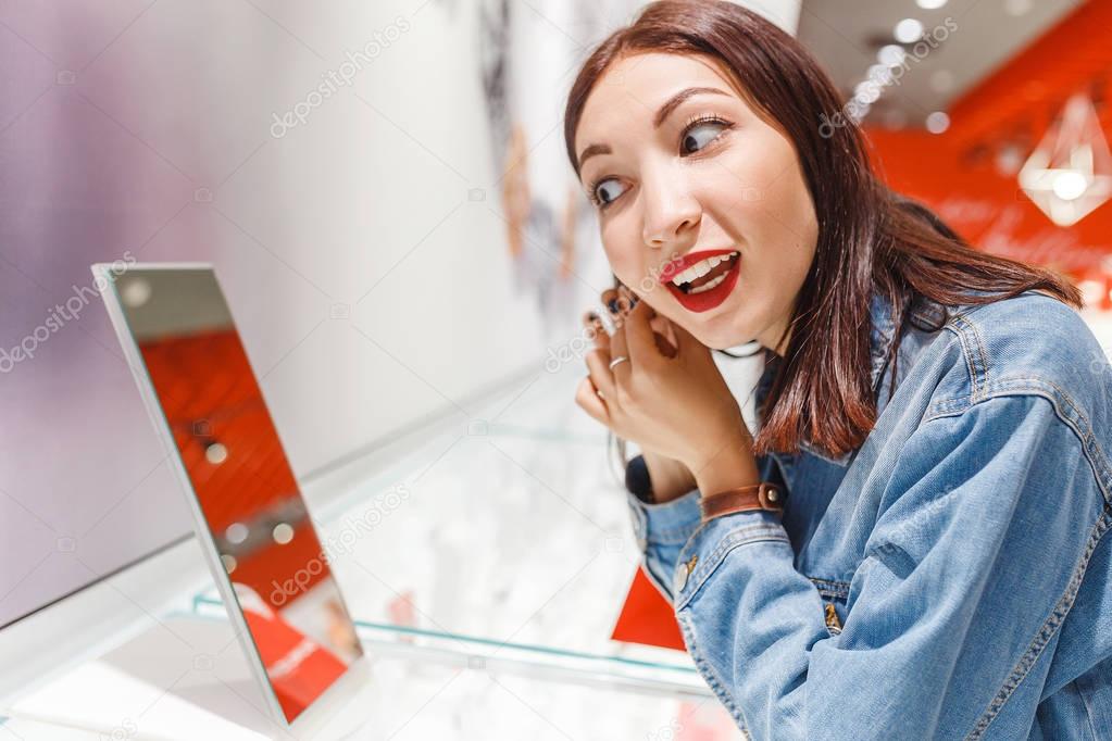 Beautiful young woman trying on earrings and looking at the mirror in jewelry store, shopping concept