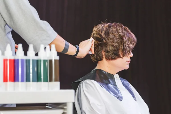 Un coiffeur styliste fait une coiffure à la cliente dans un salon de beauté sur le fond de divers produits et bouteilles pour les soins capillaires — Photo