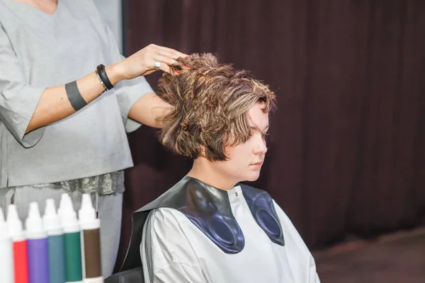 Un coiffeur styliste fait une coiffure à la cliente dans un salon de beauté sur le fond de divers produits et bouteilles pour les soins capillaires — Photo
