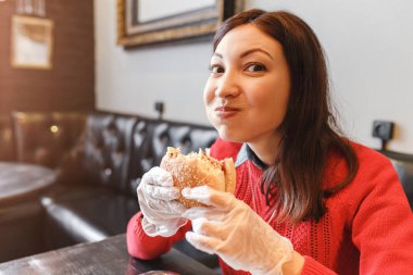 Mutlu genç kadın kapalı cafede koruyucu beyaz eldivenler, hamburger yemek