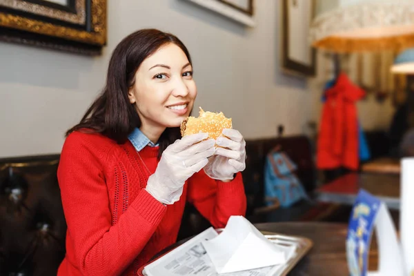Šťastná mladá žena jíst hamburger vnitřní Café v bílých rukavicích — Stock fotografie
