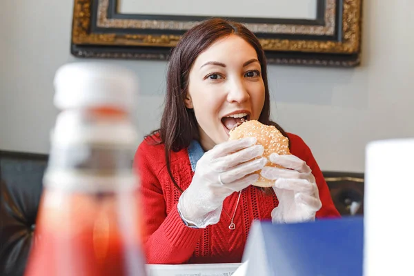 Gelukkig jonge vrouw die het eten van een hamburger in overdekte café in beschermende witte handschoenen — Stockfoto