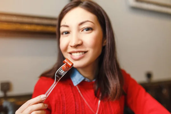 Smiling beautiful young woman eating a piece of grilled sausage using a fork in restaurant — Stock Photo, Image