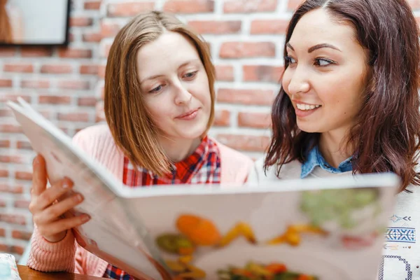 Две счастливые эмоциональные женщины сидят в современном ресторане или кафе, выбирая еду из меню и разговаривая, сплетничая. Girl Friends Leisure и концепция местной кухни — стоковое фото