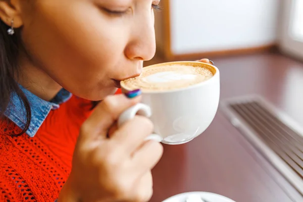 Красива молода жінка п'є каву, сидячи біля вікна в кафе — стокове фото