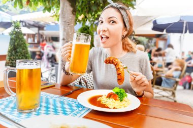 A happy Asian girl drinks a mug of lager beer in a traditional Bavarian Biergarten and snacks on a juicy sausage and potato salad. German delicacy cuisine concept clipart