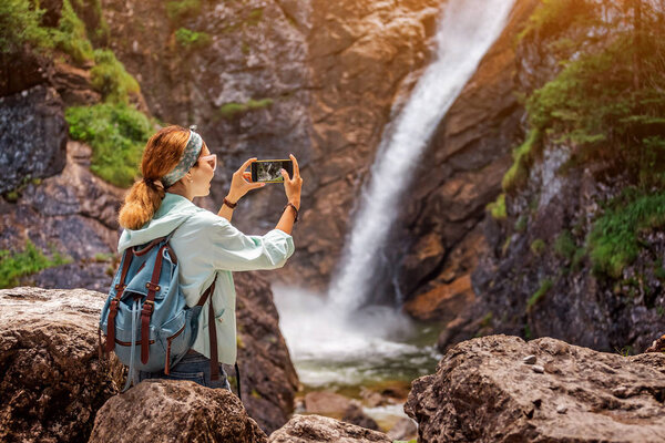Happy traveller woman taking photo of majestic waterfall in the jungle. Hiking and sightseeing concept