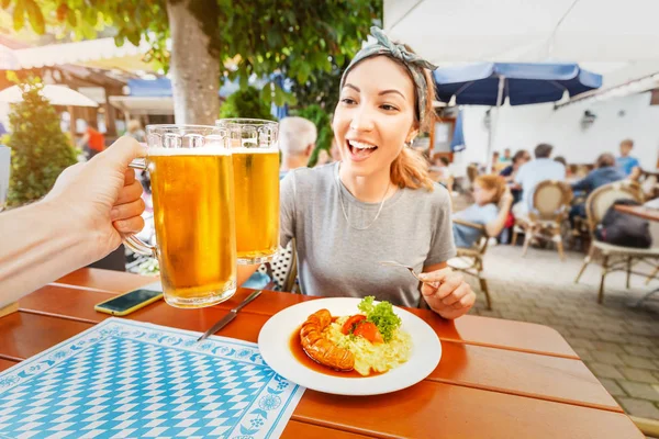 Счастливая азиатка выпивает кружку баварского пива в Биргартене. Традиционная немецкая закуска с колбасой и картофельным салатом — стоковое фото