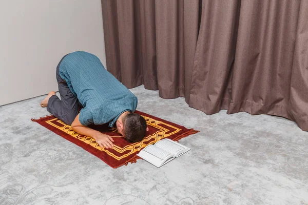 Młody człowiek modli się i recytuje muzułmański Koran na małym przenośnym dywanie we wnętrzu. Pojęcie religii i rytuału — Zdjęcie stockowe