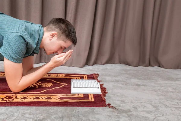 Młody człowiek modli się i recytuje muzułmański Koran na małym przenośnym dywanie we wnętrzu. Pojęcie religii i rytuału — Zdjęcie stockowe