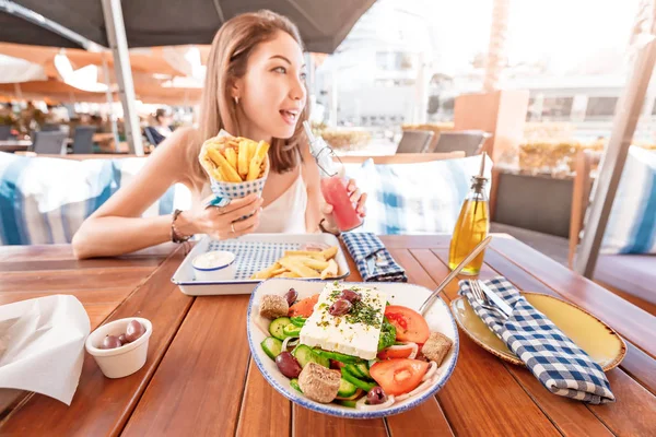 Cheerful tourist girl tries Greek cuisine at a local restaurant. On the table traditional salad Horiatiki and Gyros in Pita — Stock Photo, Image