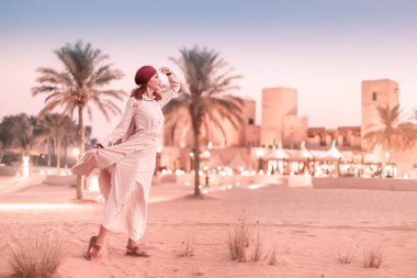 Happy asian woman in turban travels in Sahara desert. Oasis and Bedouin village at the background. Adventure and life experience concept