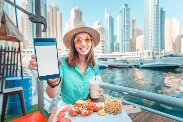 Ασιάτισσα τρώει θαλασσινά κουζίνα με θέα τους ουρανοξύστες και το λιμάνι της Μαρίνας στο Ντουμπάι και δείχνει μια κενή οθόνη του smartphone της. Έννοια της εφαρμογής για την παρακολούθηση θερμίδων και διατροφής — Φωτογραφία Αρχείου