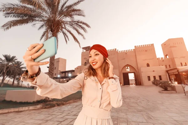 Happy woman traveler wearing dress and turban taking selfie photos on her smartphone of an old Arab town or village in the middle of the desert. Concept of tourism and adventures — 스톡 사진