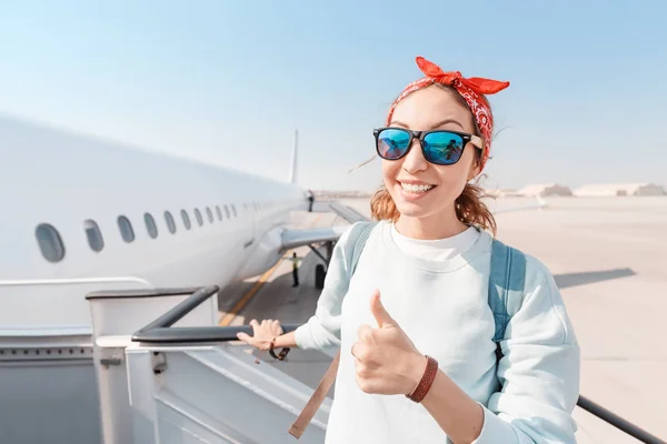 Ένα νεαρό κορίτσι χαμογελάει και σηκώνει τους αντίχειρες ενώ επιβιβάζεται σε ένα μεγάλο λευκό αεροπλάνο στο αεροδρόμιο. Έννοια του ταξιδιού και νίκη κατά της αεροφοβίας — Φωτογραφία Αρχείου