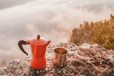 Geyser kahve makinesi ve kupası dağın tepesinde sisli şafağa bakan bir kayanın üstünde. Kamp ve açık hava eğlencesi için sofra takımı kavramı