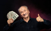 Starší pán drží hromadu peněz. Portrét vzrušeného starého muže. Šťastný starý muž drží dolarové bankovky na tmavočerveném pozadí. 