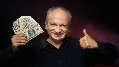 Kıdemli beyefendi bir tomar para tutuyor. Heyecanlı yaşlı bir adamın portresi. Kırmızı arka planda dolar banknotları tutan mutlu yaşlı adam.. 