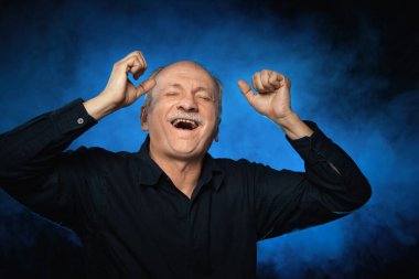 Yaşlı maço adam. Yaşlı bir adam mavi dumanlı koyu bir arka planda ellerini başının arkasında tutar. Stüdyonun karanlık arka planında yaşlı duygusal bir adamın portresi.