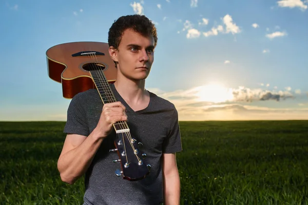 Portrait of teenager boy holding guitar and standing in field at sunset.