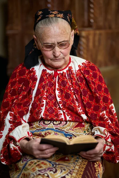 Ηλικιωμένη Γυναίκα Φορώντας Παραδοσιακή Ενδυμασία Και Γυαλιά Ανάγνωσης Βιβλίου Εσωτερική — Φωτογραφία Αρχείου
