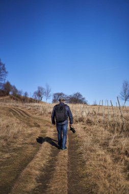 Sırt çantalı ve kameralı bir adam kırsalda yürüyüş yapıyor.