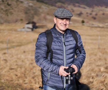 Sırt çantalı ve kameralı bir adam kırsalda yürüyüş yapıyor.