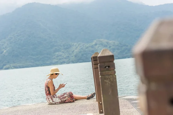 帽子をかぶった女性が携帯電話を使って桟橋に座る — ストック写真