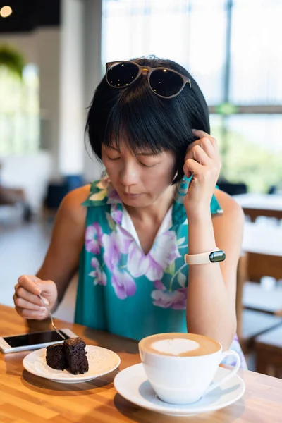 成熟したアジアの女性はコーヒーショップでチョコレートブラウニーケーキとコーヒーを食べます — ストック写真