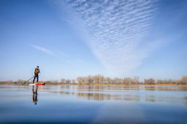 ウェットスーツのシニア男性パドラーは コロラド州 冬または早春の風景 低角度アクションカメラビュー レクリエーション フィットネス トレーニングコンセプトの湖でスタンドアップパドルボードを漕いでいます — ストック写真