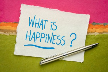 Mutluluk nedir? Renkli ve soyut manzaraya karşı küçük beyaz bir Khadi bez kağıdına el yazısı. İlham verici soru ve kişisel gelişim kavramı.