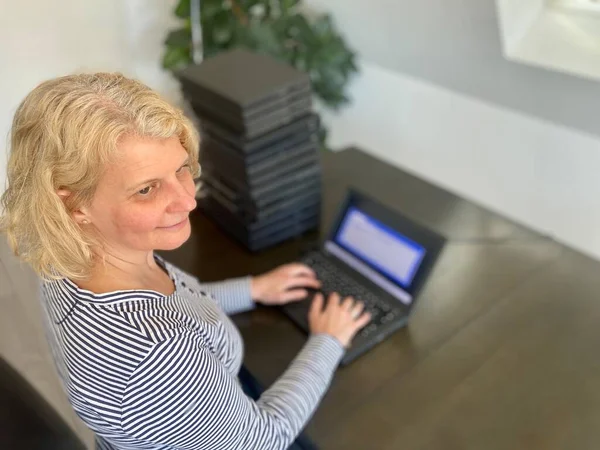 A casually dressed caucasian middle aged woman is installing a laptop computer with a stack of several more computers next to her. Laptops are used and second hand, might be for people in need due to — Stock Photo, Image