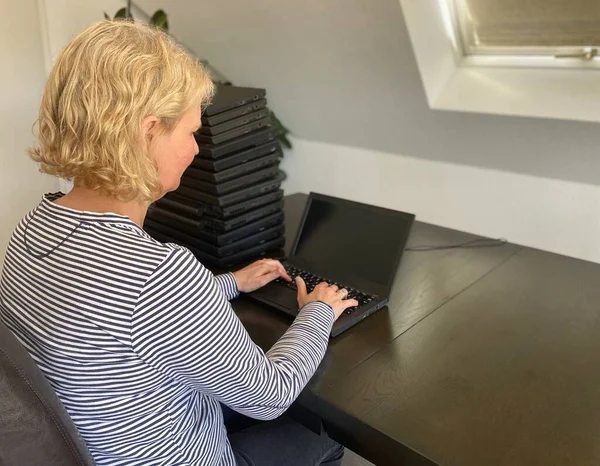 A casually dressed caucasian middle aged woman is installing a laptop computer with a stack of several more computers next to her. Laptops are used and second hand, might be for people in need due to — Stock Photo, Image