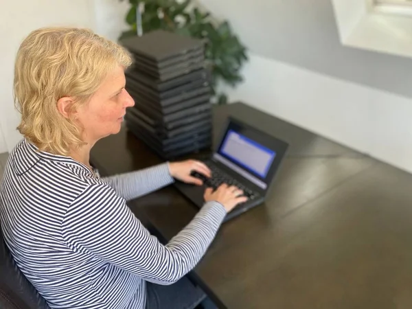 A casually dressed caucasian middle aged woman is installing a laptop computer with a stack of several more computers next to her. Laptops are used and second hand, might be for people in need due to — Stock Photo, Image