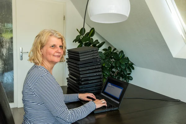 Una mujer caucásica casualmente vestida de mediana edad está instalando un ordenador portátil con una pila de varios ordenadores más a su lado. Las computadoras portátiles se utilizan y de segunda mano, podría ser para las personas necesitadas debido a — Foto de Stock