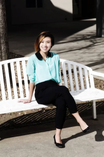 Sonriente joven asiático americana mujer sentado en banco al aire libre — Foto de Stock