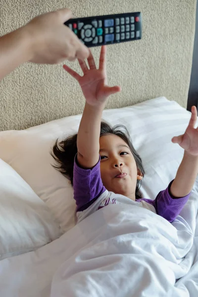 Ασιατικό παιδί στο κρεβάτι θέλει να αναλάβει τηλεόραση τηλεχειριστήριο από τα χέρια του γονέα. Εθισμός ή γονική συμβουλευτική έννοια — Φωτογραφία Αρχείου