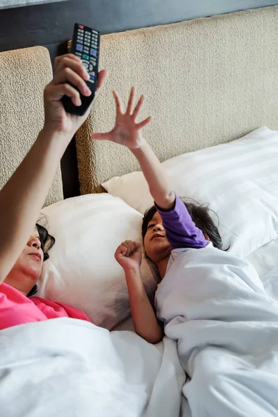 Asijské dítě na lůžku chce převzít kontrolu nad televizí dálkové ovládání z jeho rodičovských rukou. Závislost, koncepce rodičovské kontroly — Stock fotografie