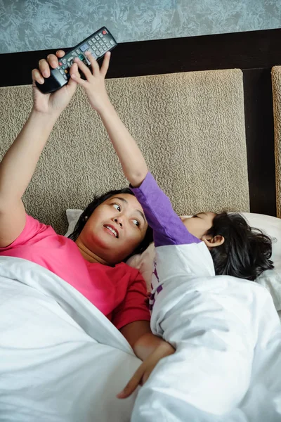 침대에 누워 있는 아시아인 아이는 어머니로부터 텔레비전 리모컨을 물려받고 싶어 한다. 부모 의지 배 스톡 사진