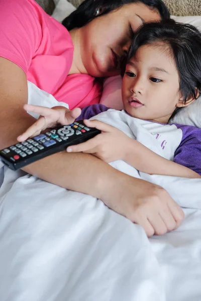 Asijské malý chlapec na lůžku pomocí dálkového ovládání sledovat televizi, zatímco jeho matka Asleep — Stock fotografie