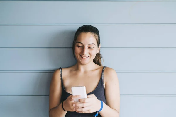 Hermosa mujer joven apoyada en una pared de madera mientras usa un teléfono inteligente — Foto de Stock