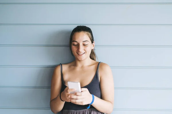 Hermosa mujer joven apoyada en una pared de madera mientras usa un teléfono inteligente — Foto de Stock