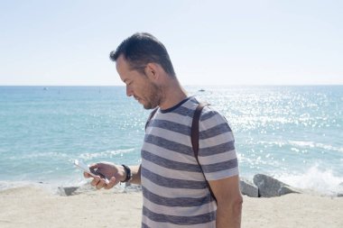 Güneşli bir günde deniz kenarında cep telefonu kullanan tişörtlü bir gencin yan görüntüsü