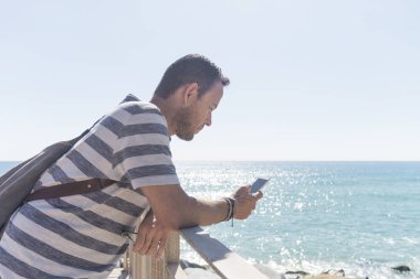 Güneşli bir günde deniz kenarında cep telefonu kullanan tişörtlü bir gencin yan görüntüsü