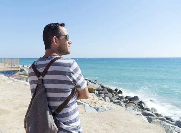 Hombre con bolso de hombro de pie en el paseo marítimo mirando al horizonte en un día soleado — Foto de Stock
